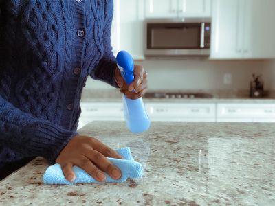 A person cleans a kitchen counter with a spray bottle of cleaner and a cloth