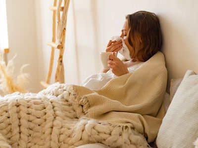 Sick young woman lying in bed covered with blanket holding a cup of hot tea