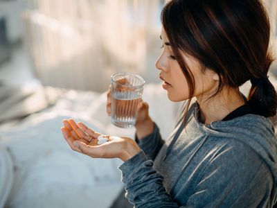 Young Asian woman sitting on bed and feeling sick, taking medicines in hand with a glass of water