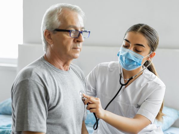 An older White male with glasses having his heart listened to by a young provider with long hair and a face mask who is using a stethoscope.