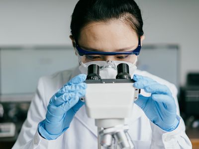 person wearing goggles and gloves looking into a microscope in a lab