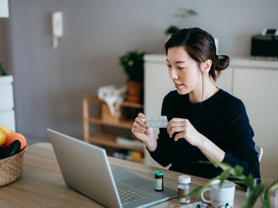 Young Asian woman video conferencing with laptop to connect with her family doctor, consulting about medicine during self isolation at home in Covid-19 health crisis