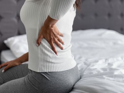 Woman with side back pain sitting on bed at home - stock photo