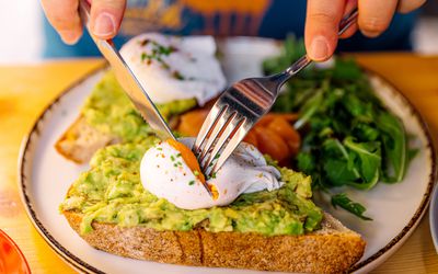 Close up image of hands holding a fork and knife, cutting into a paoched egg atop avocado toast with a side of arugula. 