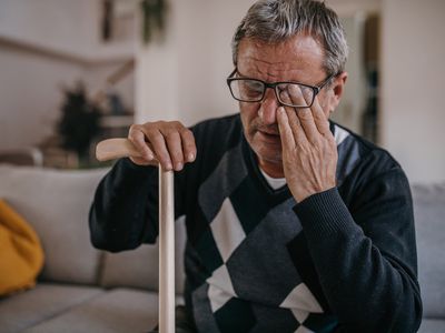 Older man with cane sits on couch holding his eye with his hand