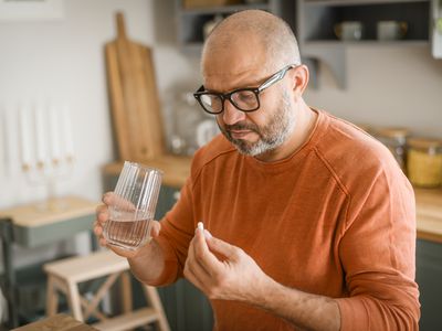 Man in orange shirt and thick rimmed glasses taking a supplement with a glass of water, while standing in a kitchen.