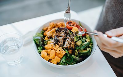 Close up, high angle shot of young woman enjoying multi-coloured healthy fruit, vegetables with grilled chicken salad bowl with balanced nutrition in cafe, with a glass of water by the side.