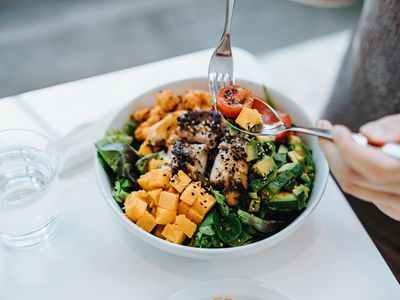 Close up, high angle shot of young woman enjoying multi-coloured healthy fruit, vegetables with grilled chicken salad bowl with balanced nutrition in cafe, with a glass of water by the side.