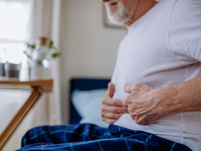 Unrecognizable senior man, sitting on the edge of a bed clutching his stomach