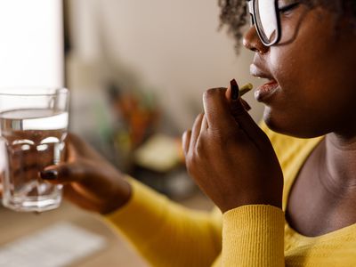 Close up shot of young woman sitting at her desk at the office and putting a pill in her mouth when taking medicine at work.