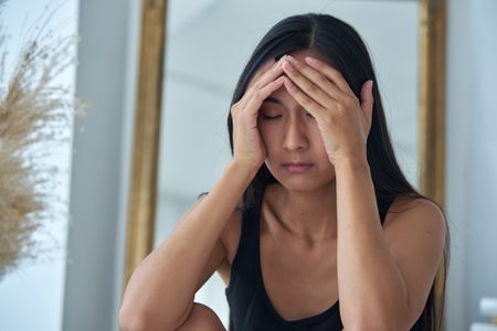 Stressed anxious Asian woman holding head in hands, keeping eyes closed, suffering from headache or migraine. Stress and anxiety concept