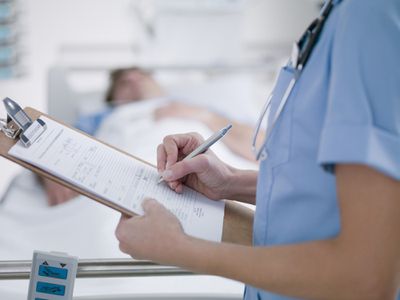 nurse writing on clipboard and man in hospital bed in the background