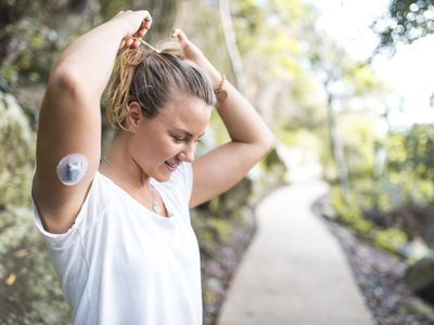 A White adult tying back their blonde hair with their arm raised, you can see they have a CGM sticker on.