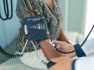 A mature adult woman is at a routine medical check. She is sitting on an examination table in a clinic. The nurse is using a blood pressure gauge to check the patient's blood pressure.