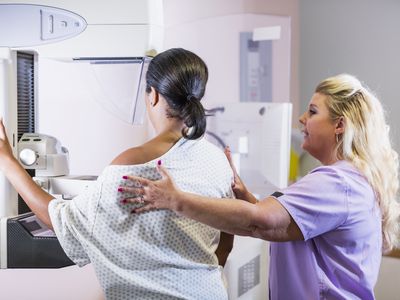 A mature African-American woman in her 40s wearing a hospital gown, getting her annual mammogram. She is being helped by a technologist, a blond woman wearing scrubs.