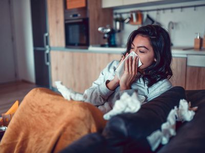 Female at home sick, sneezing into a tissue while sitting on the couch