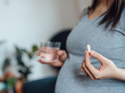 Close up of pregnant person holding a pill in one hand and a glass of water in the other