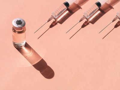 Plastic disposable sterile syringe and sealed vial of liquid remedy placed on pastel peach orange background with shadows and light reflection