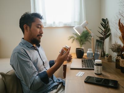 Man looking at pill bottle while sitting at desk and listening to a doctor on a video conference at home