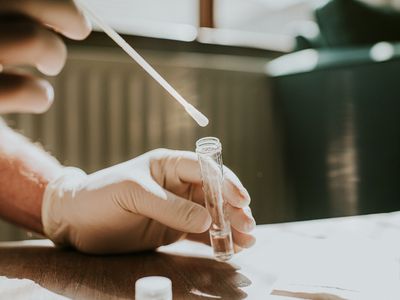 A hand wearing a latex glove holds a small clear tube and dips a cotton swab into the liquid