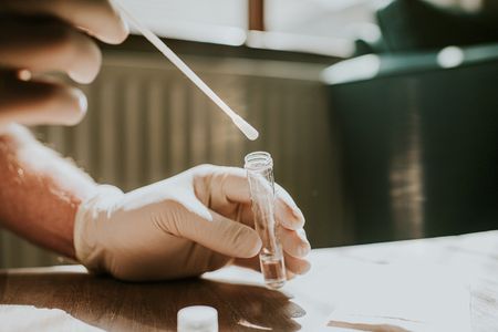 A hand wearing a latex glove holds a small clear tube and dips a cotton swab into the liquid