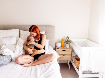 Woman holding newborn baby while sitting on bed next to a breastfeeding pillow