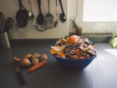 vegetable scraps in a container with a knife and unpeeled vegetables next to it