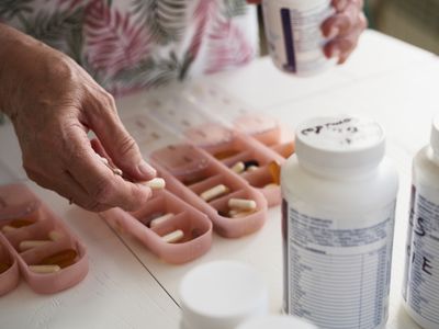 Older woman's hand filling weekly pill organizer with capsules while pill bottles sit on white table