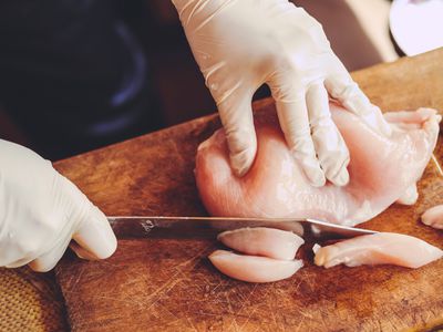 Gloved hands slicing raw chicken fillet on a wooden cutting board