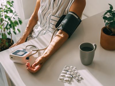 An older woman takes her blood pressure at home beside a blister pack of pills and a mug on the table