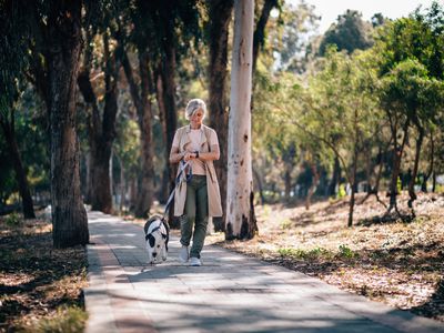 A person walks outdoors on path through park with dog on leash