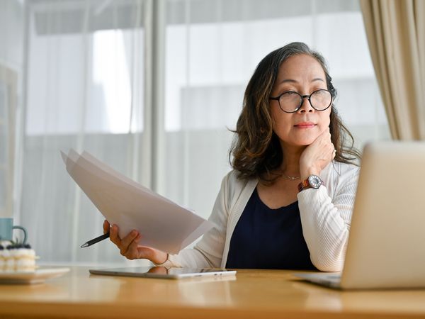 Woman works at desk with laptop, holding papers and pen